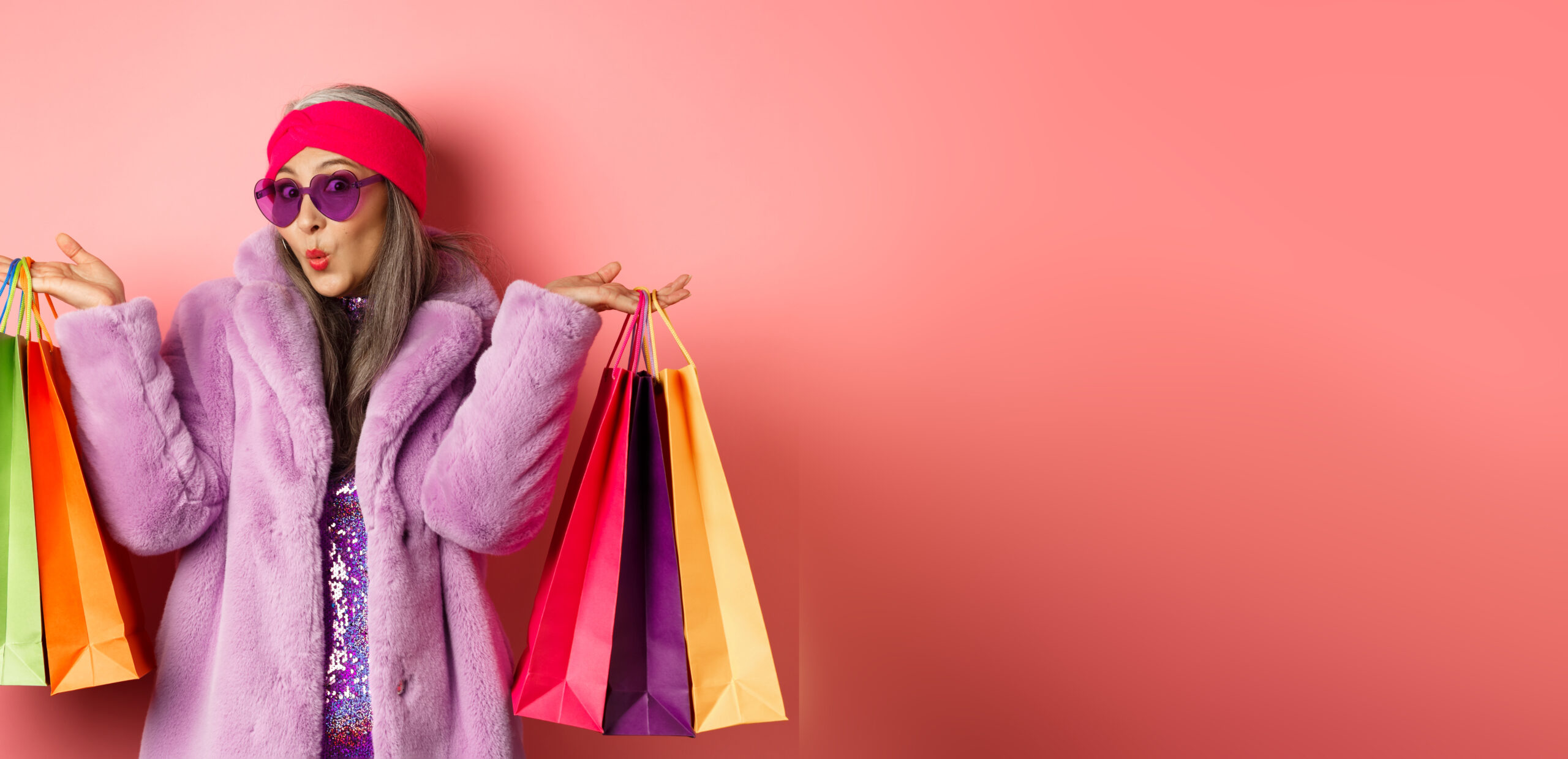 Stylish asian senior woman going shopping, wearing trendy clothes and sunglasses, holding store bags with clothes and purchases, pink background.