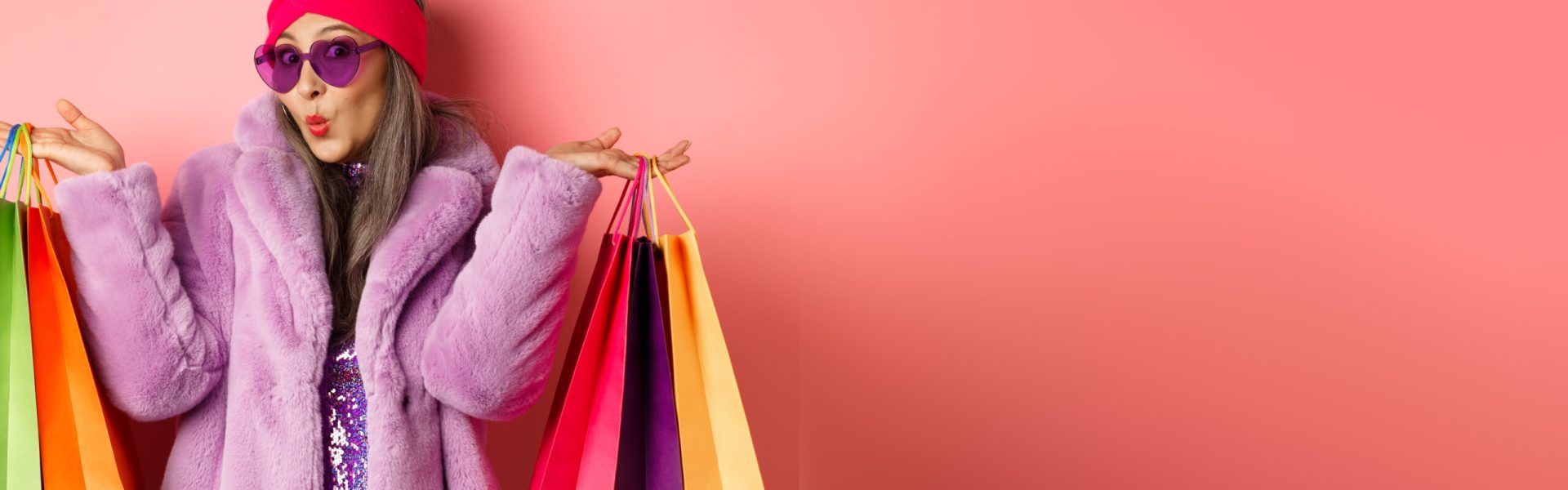 Stylish asian senior woman going shopping, wearing trendy clothes and sunglasses, holding store bags with clothes and purchases, pink background.